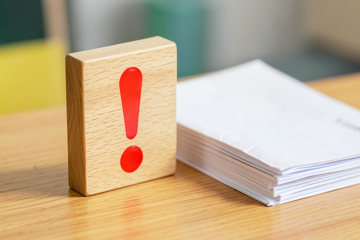 A small wooden sign with an exclamation mark, on a desk next to a stack of documents, symbolizing important information.